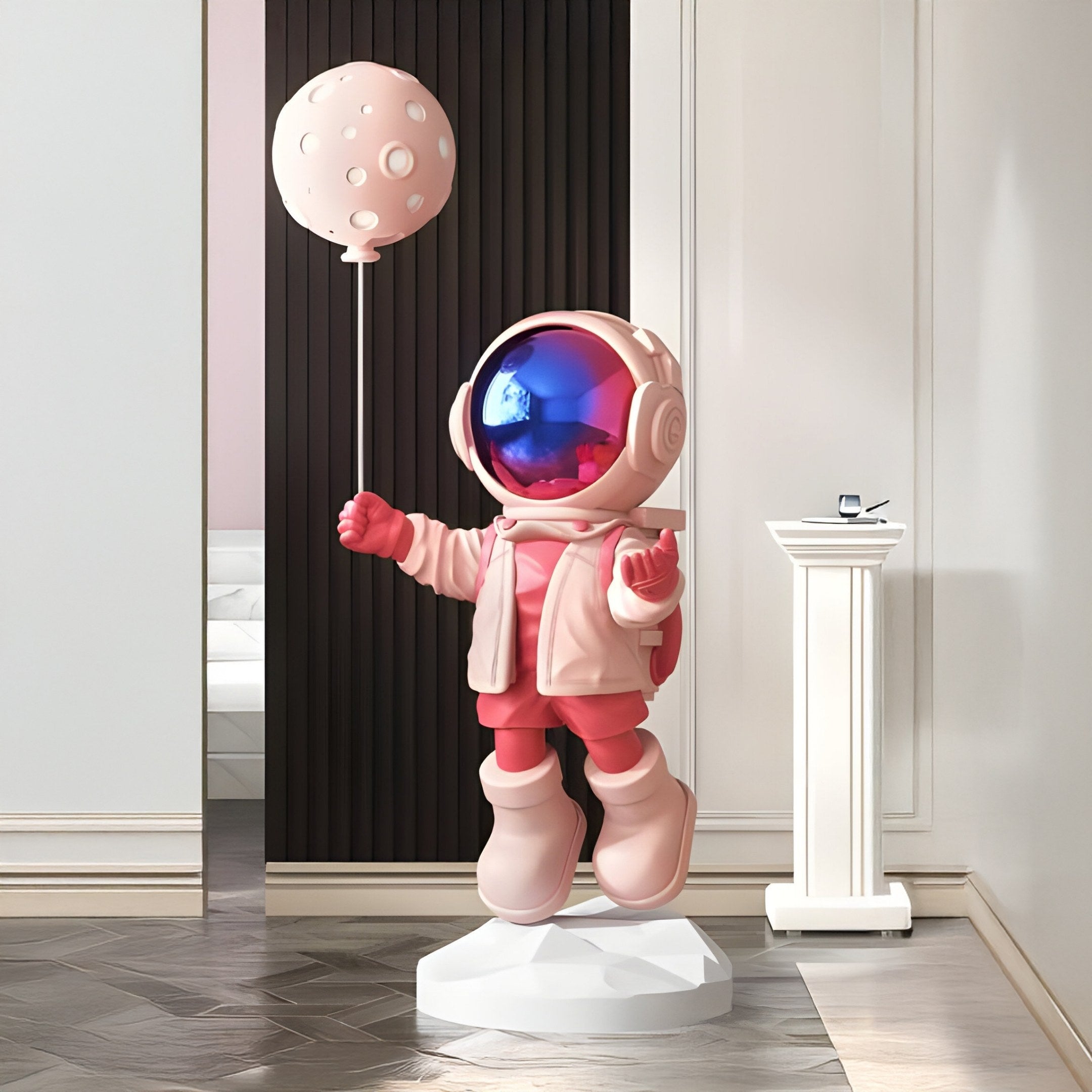 The Stellar Pink Astronaut Moon Balloon Sculpture - 160cm by Giant Sculptures features an astronaut in pink and white holding a planet-shaped balloon, adding a cute celestial touch to any modern, minimalistic room. It stands large with a small pedestal in the background.