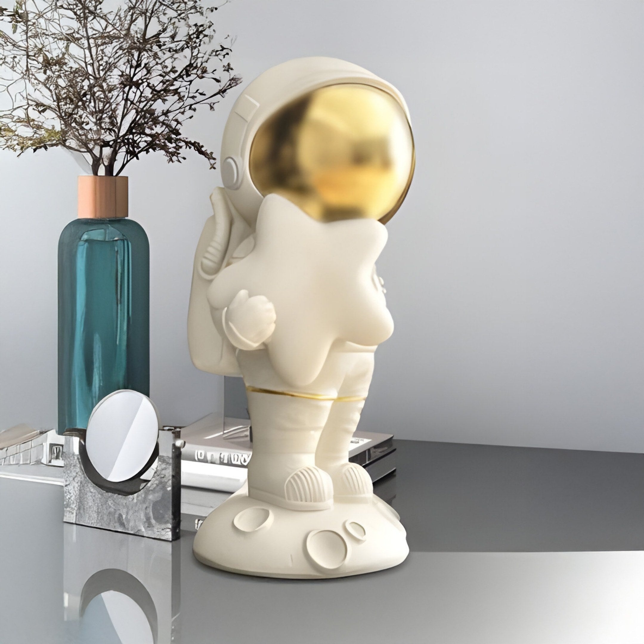 A Cloud White Stardust Moon Astronaut Figure from Giant Sculptures, 20cm tall with a shiny gold visor holding a star, sits on a gray surface beside a teal vase with dried flowers and a round mirror against a light gray wall.