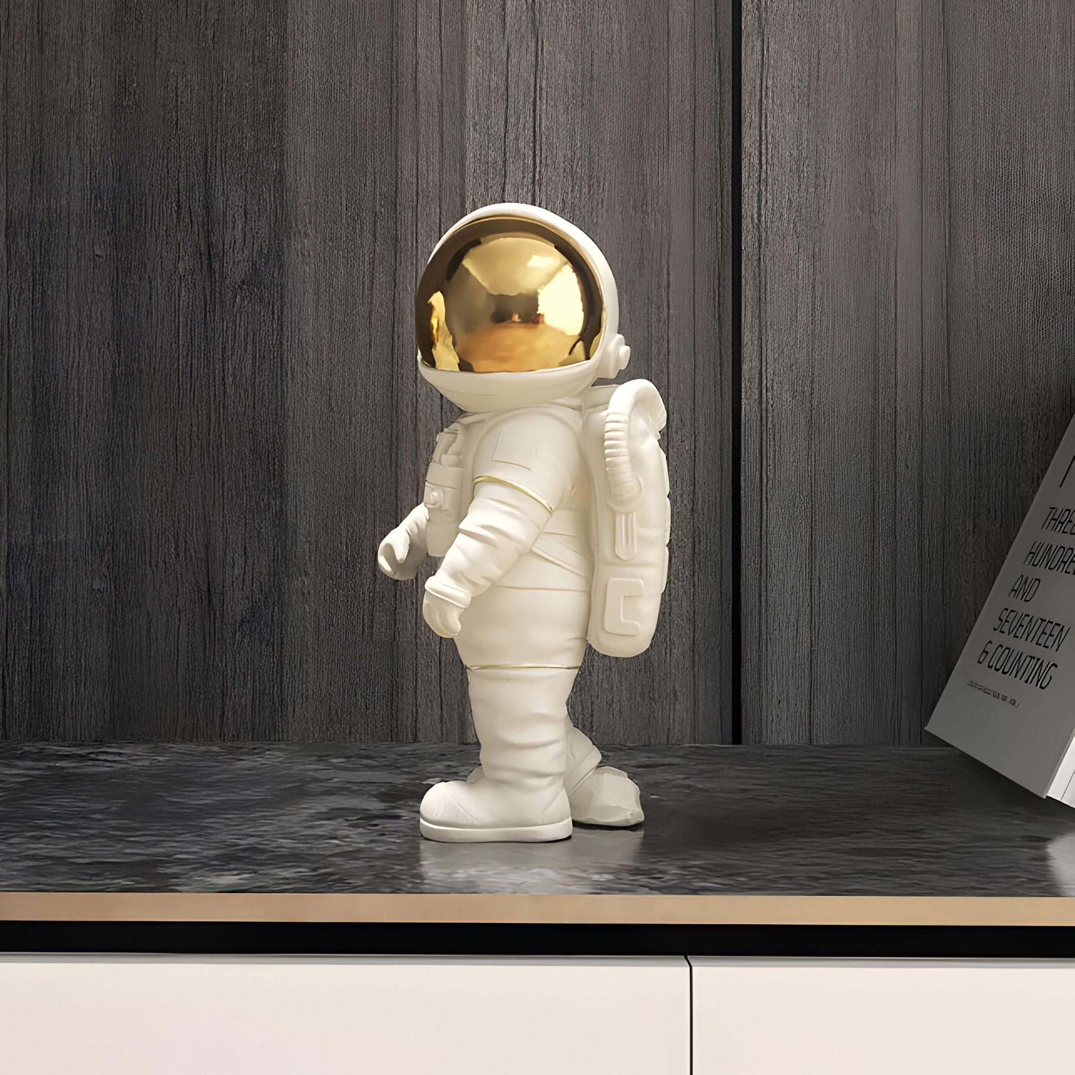 The Cloud White Stardust Astronaut Figure by Giant Sculptures, standing 20cm tall in a white suit with a reflective gold visor, embodies the spirit of space exploration on a wooden shelf against a textured dark wall and framed art, adding depth to its display.