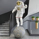 White Space Astronaut Moon Explorer Sculpture by Giant Sculptures, 200cm resin figure in white suit with gold visor standing on a grey cratered moon base, displayed beside a modern staircase with grey walls and gold hanging planters.