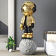 Celestial Gold Astronaut Beats On Moon Sculpture by Giant Sculptures, 210cm resin figure in gold jacket and black suit with reflective gold visor and headphones, standing on a grey cratered moon base, displayed in a modern living space.