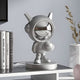 The Cosmic Chrome Astronaut DJ Figure - 30cm by Giant Sculptures stands on a tabletop with a shiny silver finish, round helmet, and reflective visor. It strikes a playful pose with one hand on its hip, perfectly complementing the surrounding books and dried grasses in the minimalist setting.