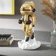 The Giant Sculptures Celestial Gold Astronaut Lift-Off Mini Figure (30cm) features a shiny backpack and floats on a white cloud atop a gray surface, displayed next to books, a glass vase with dried grass, and a tray.