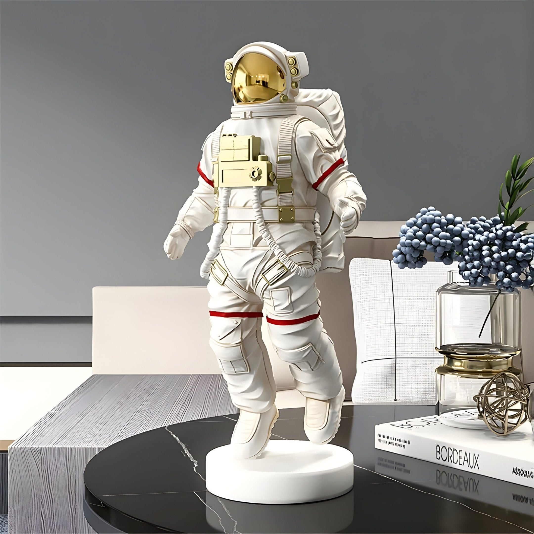 A Giant Sculptures Cloud White Space Astronaut Voyager Figure with gold details stands on a round base. The rooms neutral walls enhance the space-themed décor, with a book titled Bordeaux and a glass container holding blue flowers on the nearby table.