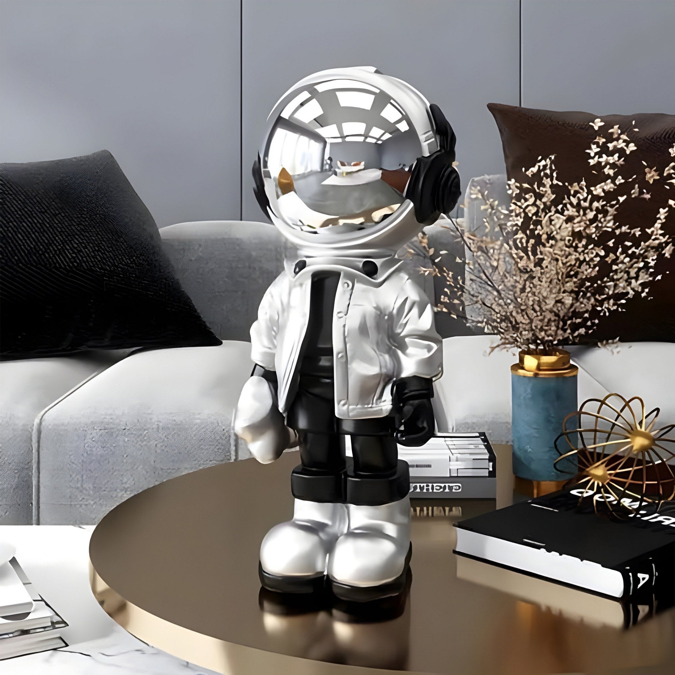 The Lunar Silver Astronaut Beats Figure by Giant Sculptures stands on a round coffee table amid books, decor, and a vase of dried flowers, set against a gray couch with black and brown cushions.