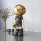 The Giant Sculptures Midnight Black Astronaut Buddy Figure - 36cm features a sleek resin design, gold helmet, and stands on a reflective surface. Its cosmic décor perfection, paired with dried branches in a glass vase and a small blue container against a plain wall backdrop.