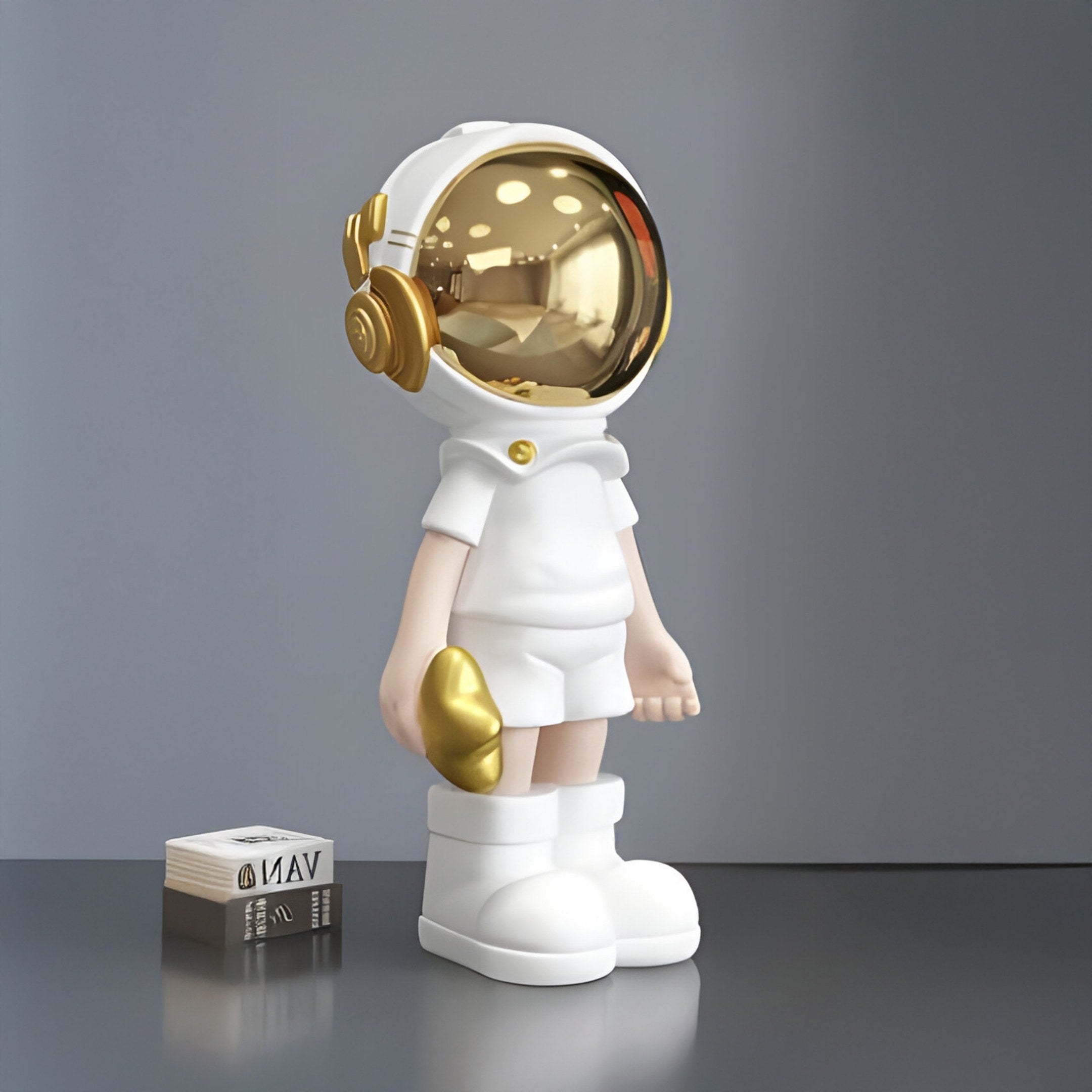 A 36cm Cosmic White Astronaut Buddy Figure by Giant Sculptures, with a reflective gold helmet, stands on a gray surface. Nearby is a small white and black box labeled Van, enhancing the space-themed décor against the plain background with subtle reflections.