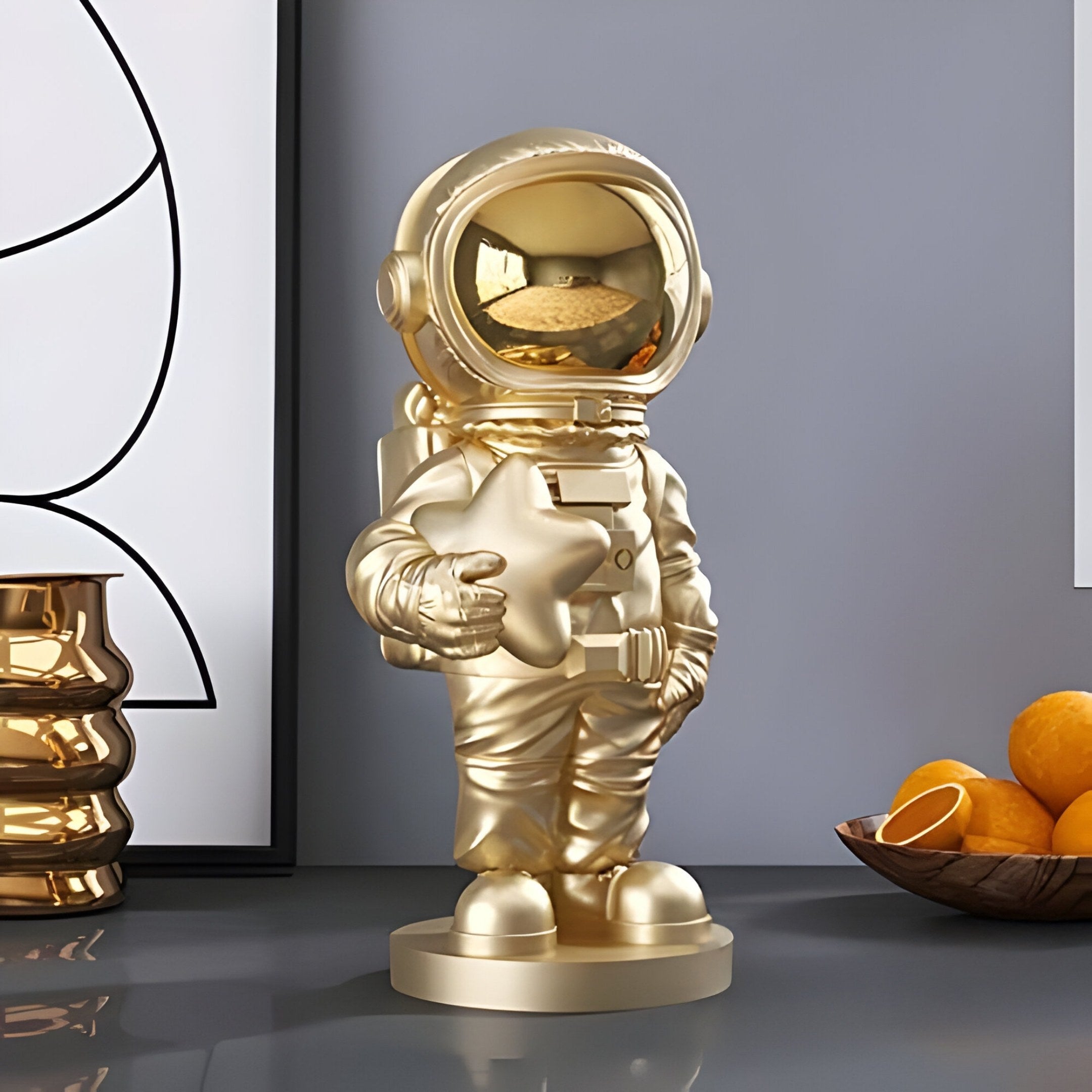 A 37cm Celestial Gold Astronaut Star Figure by Giant Sculptures is placed on a surface with a modern abstract painting. Nearby, a shiny gold vase and bowl of oranges enhance the imaginative décor, adding cosmic accents to the contemporary setting.