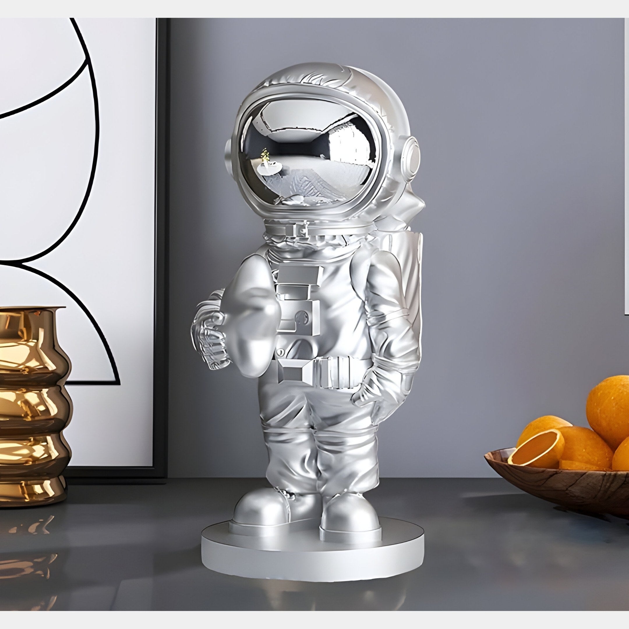 The 37cm Lunar Silver Astronaut Star Figure by Giant Sculptures, with its reflective helmet, enhances the space-themed ambiance, as golden abstract vases and a bowl of oranges offer contrast. Nearby, black and white artwork further complements the cosmic charm.