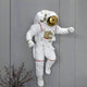 The White 3D Cosmic Lift Astronaut Wall Art by Giant Sculptures features a resin astronaut with a gold-tinted visor on a gray wall. A small bush peeks from below, while red and gold suit accents enhance its charm, creating an illusion of floating—a perfect space decor piece.