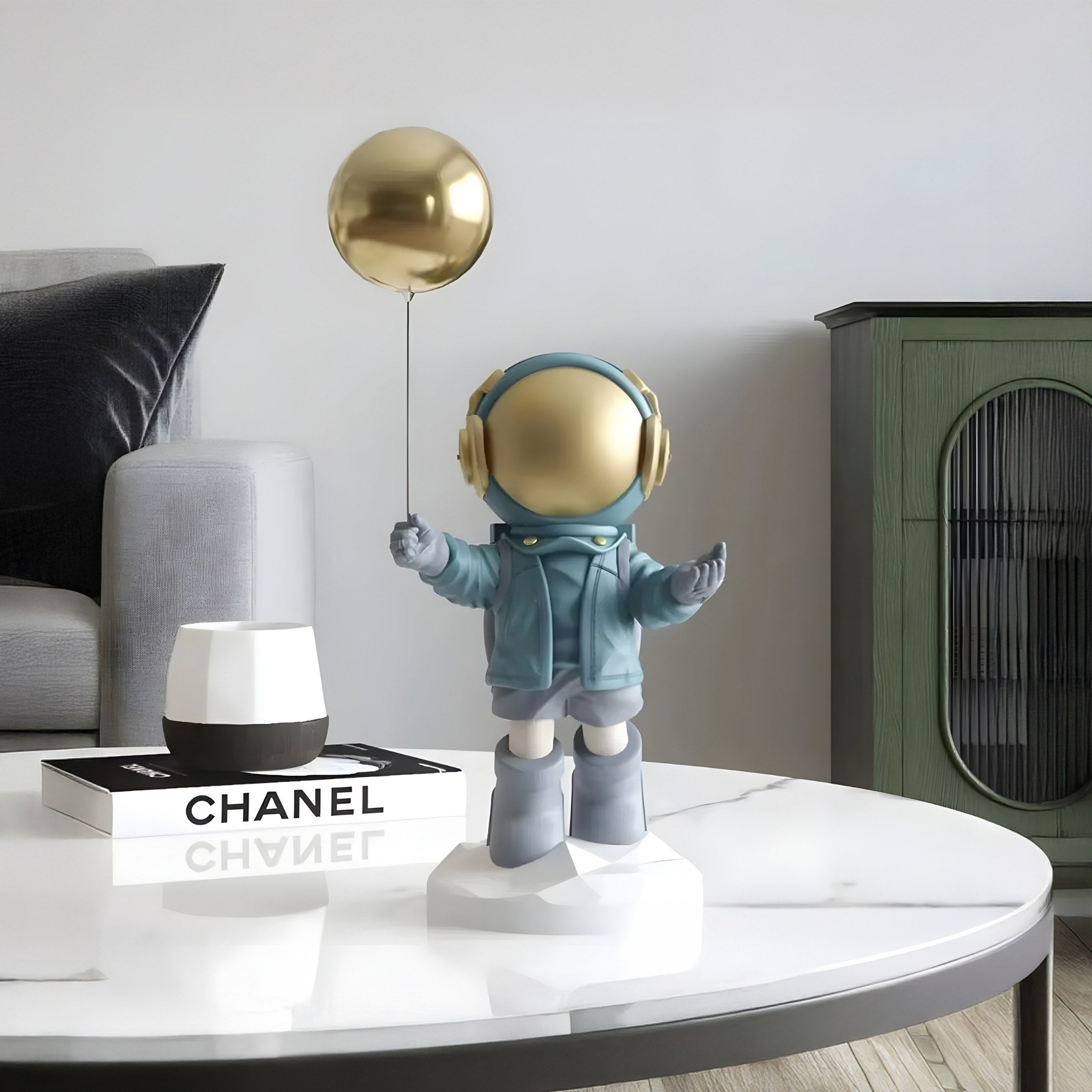 A 42cm Galactic Blue Astronaut Balloon Figure by Giant Sculptures features a gold helmet and headphones, holding a gold balloon on a round marble table. Nearby, a white candle on a Chanel book, plus a gray chair and green cabinet enhance the space-themed displays charm.