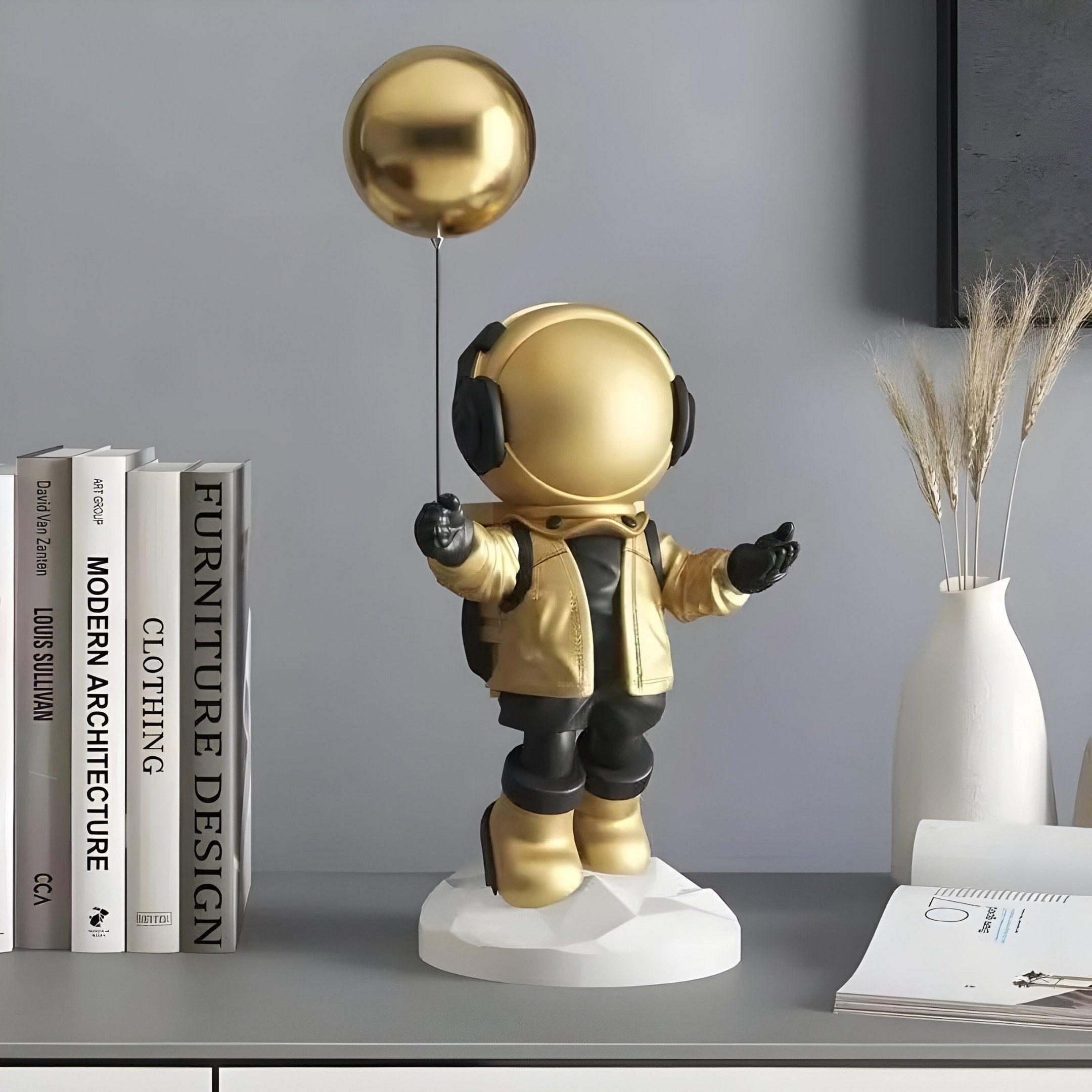 The 42cm Celestial Gold Astronaut Balloon Figure by Giant Sculptures, with black details on a white base, graces a shelf amid architecture books and a vase of dried grasses, ideal for indoor décor.