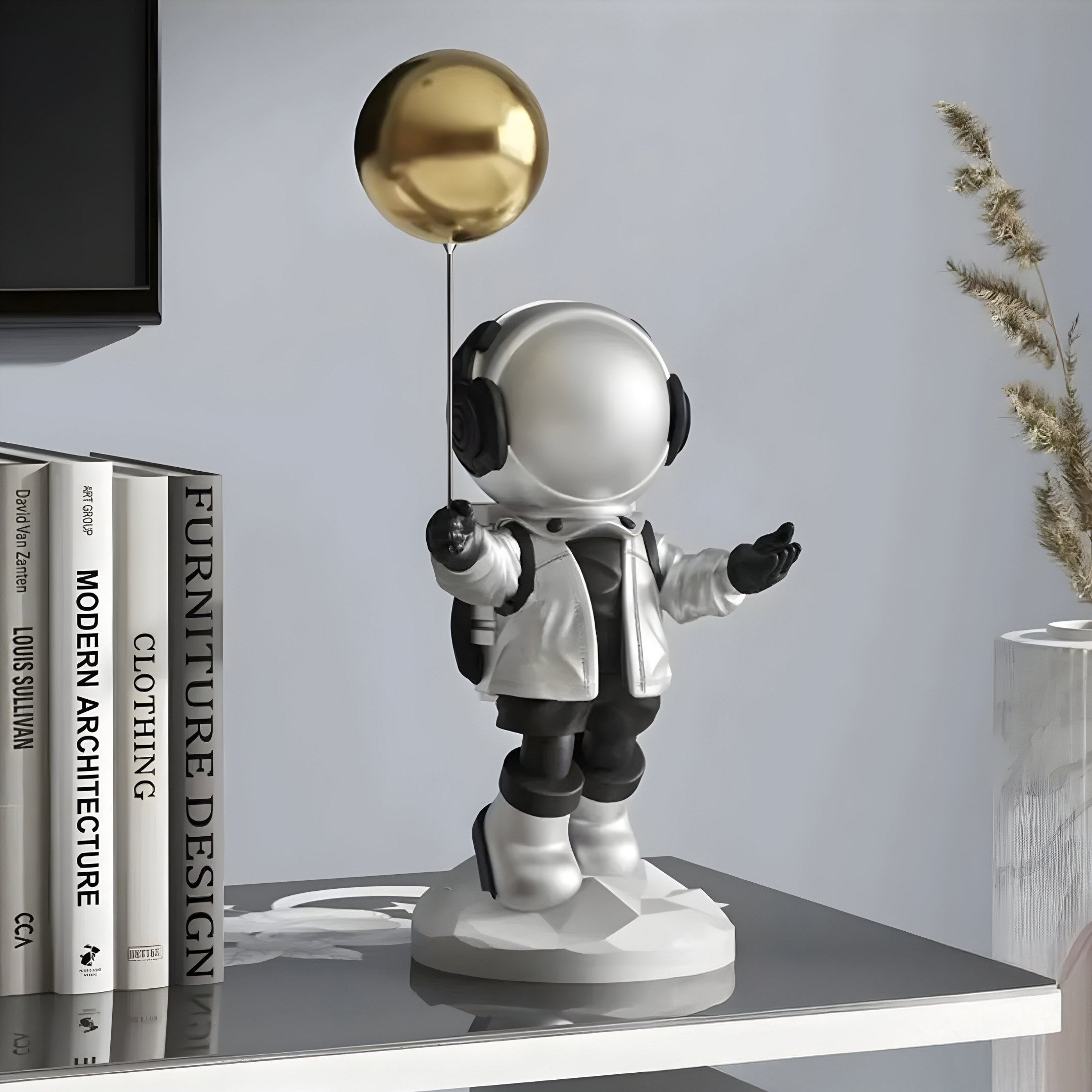 A Giant Sculptures 42cm Lunar Silver Astronaut Balloon Figure adorns the table while Modern Architecture and Furniture Design books are stacked nearby. The decor includes a plant against a gray wall with white trim.