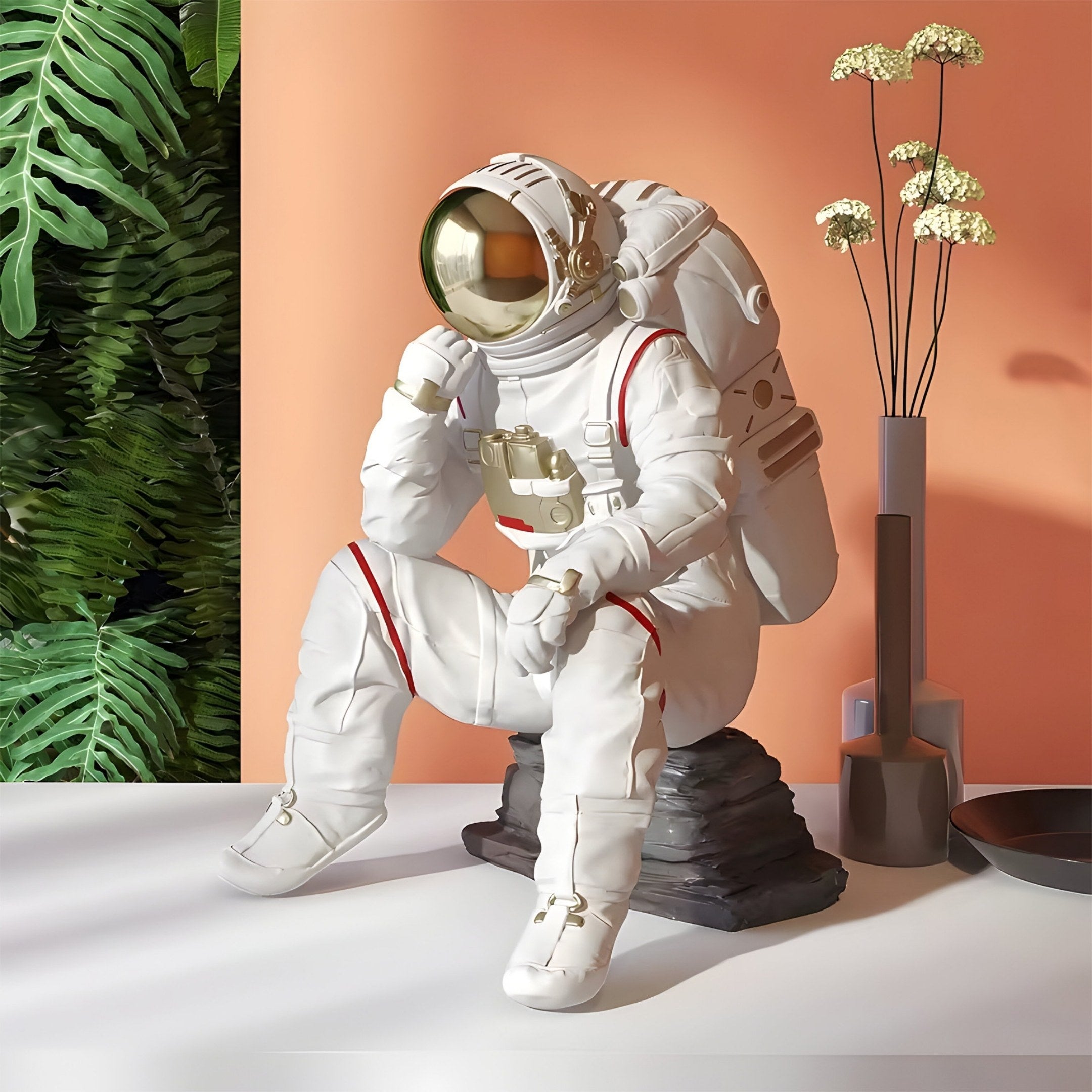 The 43cm Nova White Astronaut Thinker Figure by Giant Sculptures features a reflective white spacesuit and sits on a rock against an interstellar-themed peach background with tall plants and a flower-filled decorative vase.