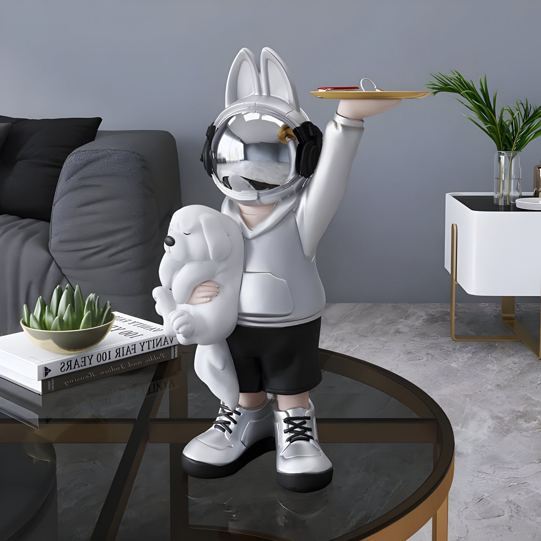 The Silver Astronaut Bunny Pup Butler Figure by Giant Sculptures, standing 44cm tall, showcases a futuristic design with a reflective helmet, hoodie, and headphones while holding a small bear. Its set against a cozy couch, lush plant, and books for a modern decor ambiance.