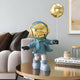 A Galactic Blue Astronaut Balloon Standing Figure (48cm) by Giant Sculptures adds a modern touch to the table. The room, featuring a beige and white couch, throw pillows, candles, and framed artwork, creates a stylish vibe.