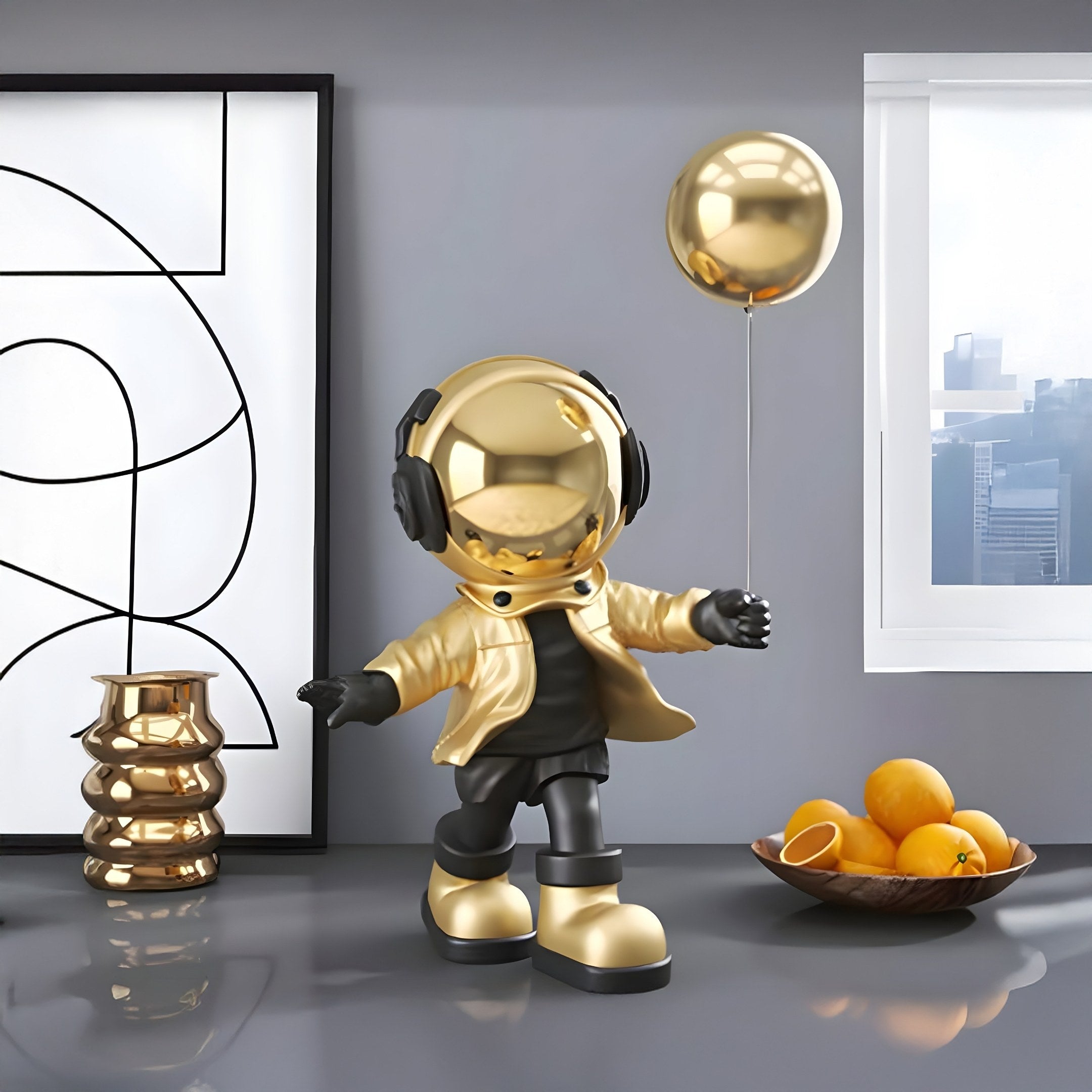 A Celestial Gold Astronaut Balloon Standing Figure by Giant Sculptures, 48cm, with a shiny helmet and jacket, adorns a modern table near a window, alongside a bowl of oranges and abstract wall art in the background.