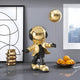 A Celestial Gold Astronaut Balloon Standing Figure by Giant Sculptures, 48cm, with a shiny helmet and jacket, adorns a modern table near a window, alongside a bowl of oranges and abstract wall art in the background.