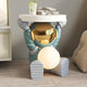 The Galactic Blue Astronaut Glow Side Table Figure (50cm) by Giant Sculptures stands on a wooden floor, holding a round top with its gold reflective helmet and glowing orb, creating ambient light. A drink and glasses sit on the modern tabletop.