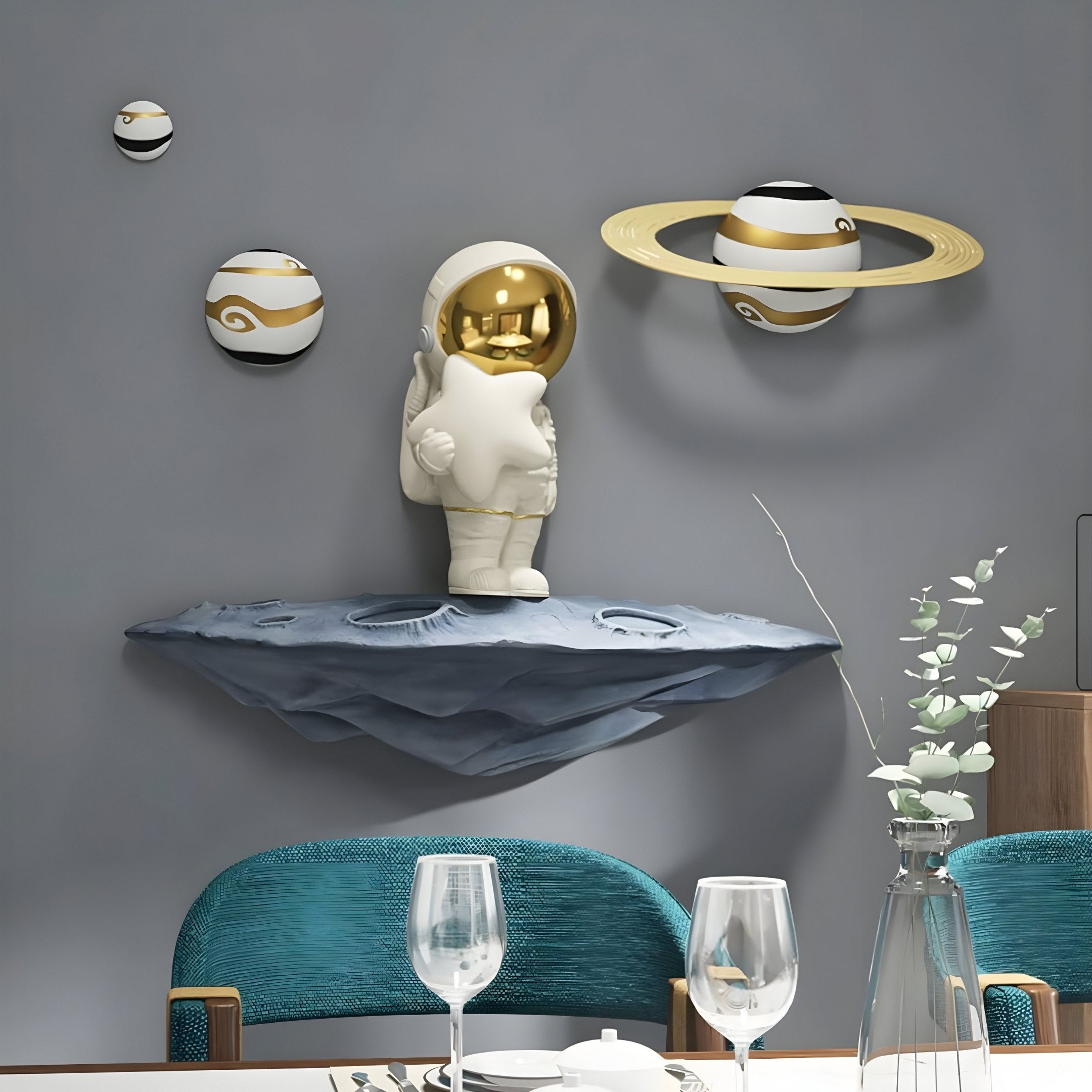 The White & Gold Orbit Odyssey Star Astronaut 3D Wall Art by Giant Sculptures features a small astronaut holding a star on a moon-like ledge, with three celestial spheres, including one resembling Saturn, enhancing the scene above a dining table with teal chairs and glassware.