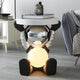 The Lunar Silver Astronaut Glow Side Table Figure (50cm) by Giant Sculptures, featuring a glowing belly, sits beside a gray armchair and white cabinet in a contemporary living room.