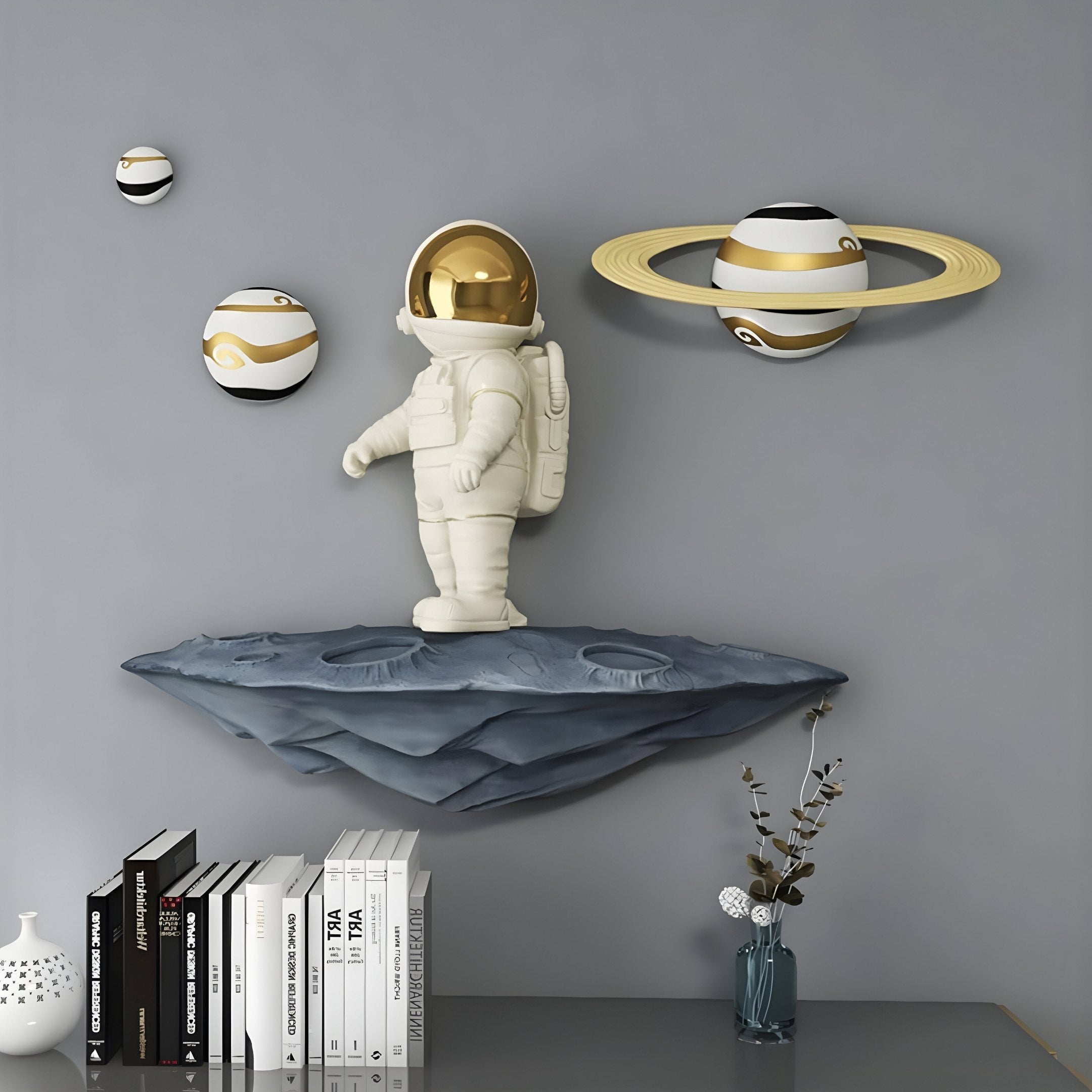 The White & Gold Orbit Odyssey Astronaut 3D Wall Art by Giant Sculptures features an astronaut figurine on a floating platform with three planetary spheres, including one with rings. Beneath, a shelf holds books and a vase with a plant—ideal for space exploration fans.