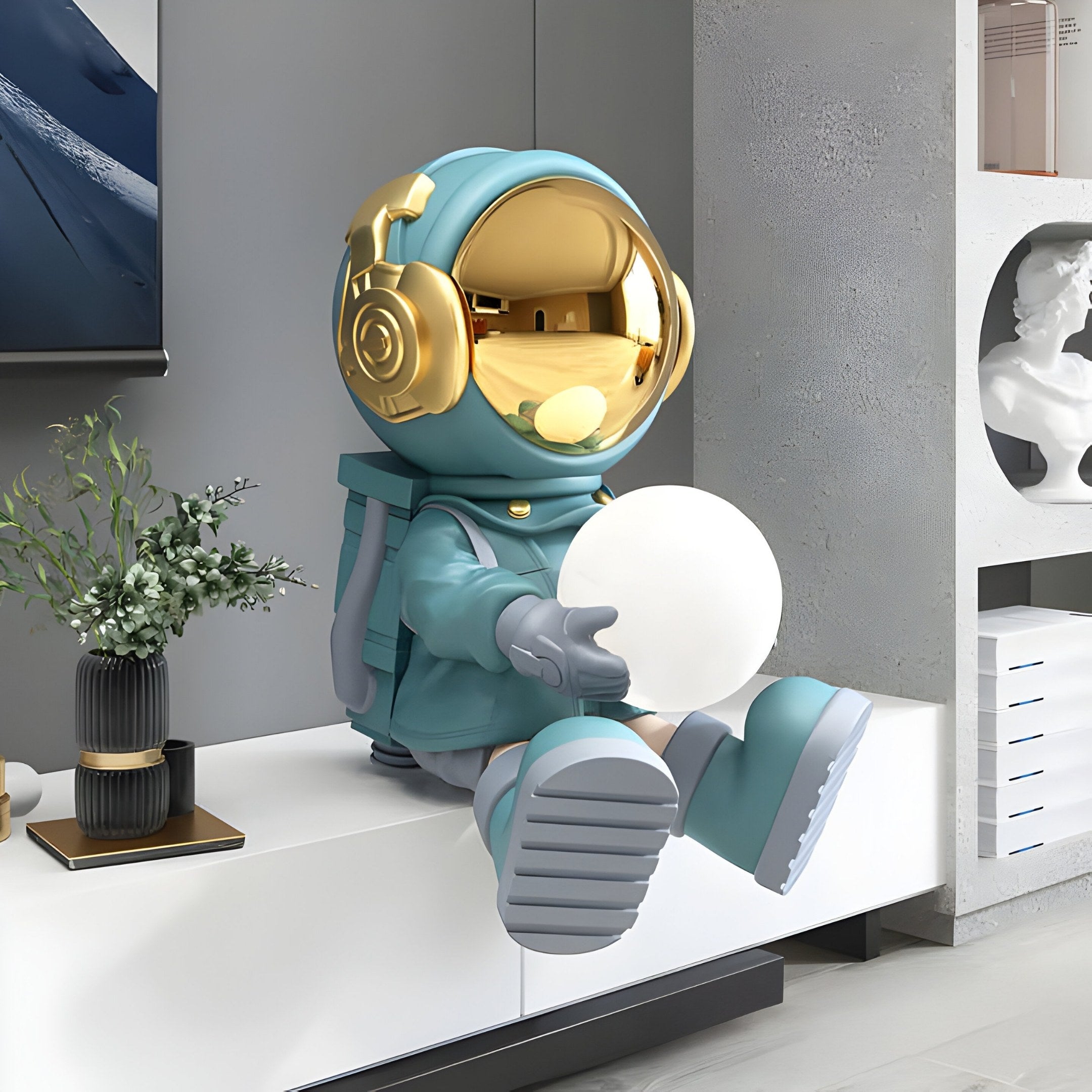 The Giant Sculptures Galactic Blue Astronaut Glow Seated II Sculpture - 53cm features a shiny gold helmet, teal suit, gray boots, and a glowing white orb with four lighting colors. It sits on a modern white shelf amidst plants and artistic decor.