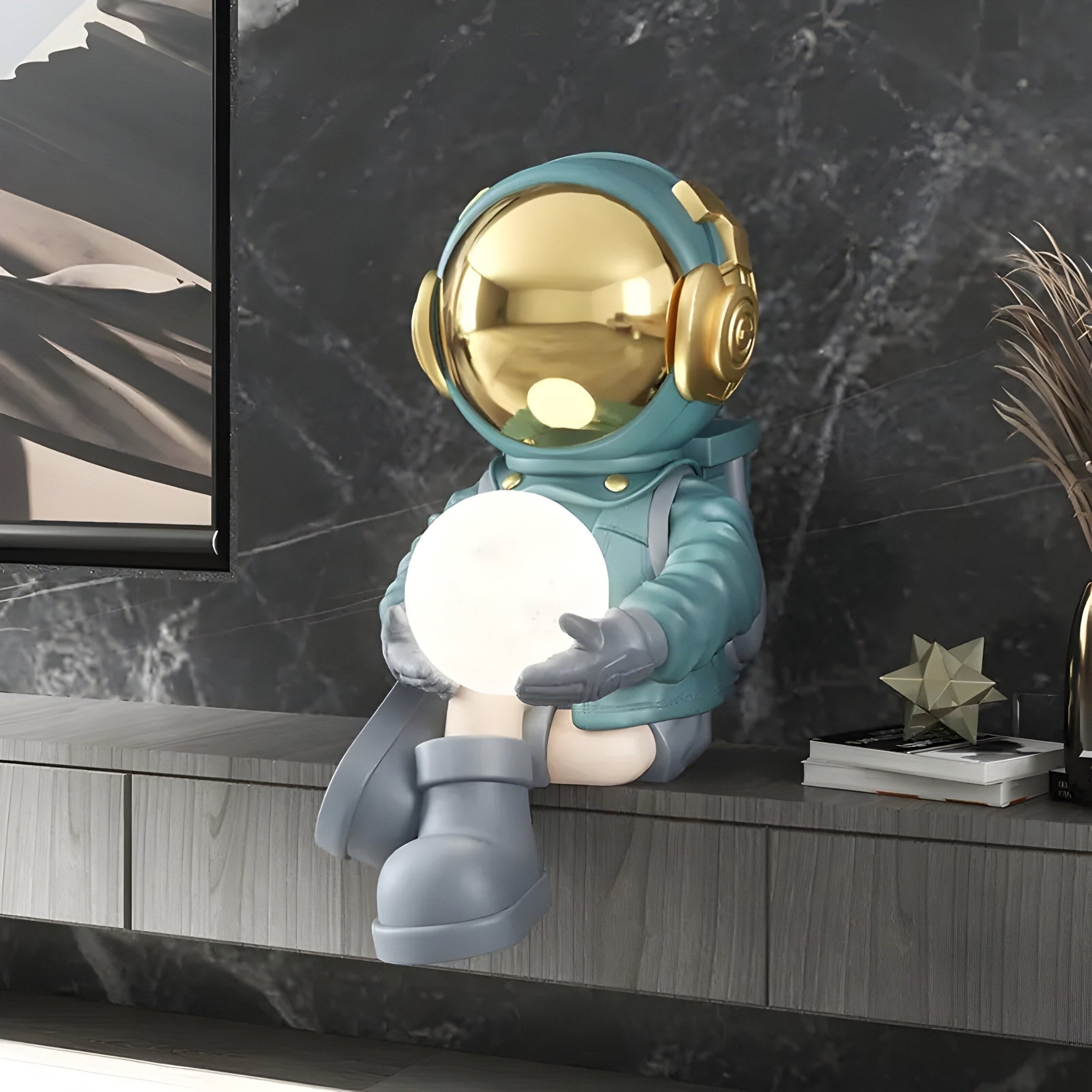 Giant Sculptures Galactic Blue Astronaut Glow Seated I Sculpture features a shiny gold helmet, cradles a glowing orb, and is 53cm tall. This battery-powered lamp offers adjustable lighting colors on a dark marbled shelf for an out-of-this-world ambiance.