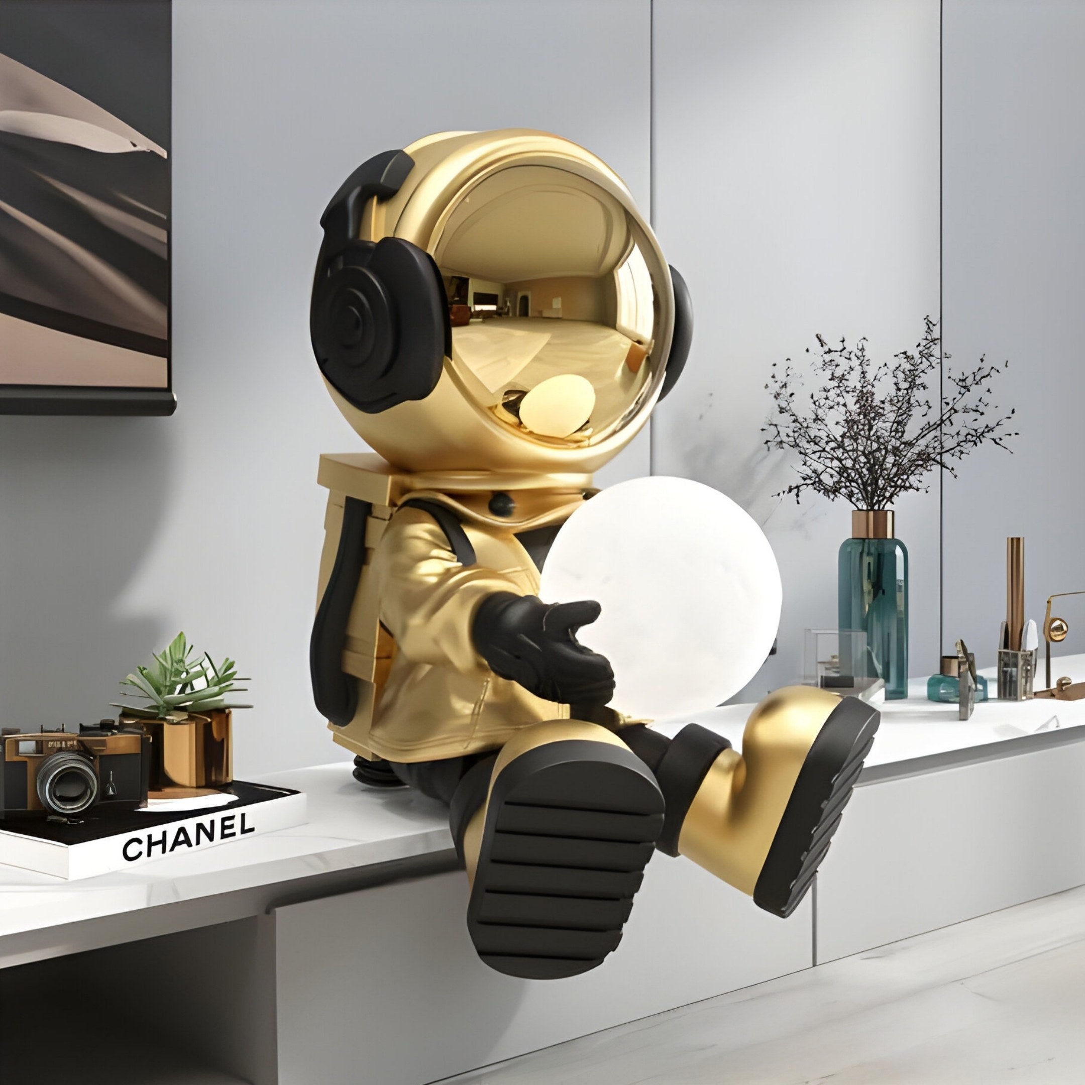 The 53cm Celestial Gold Astronaut Glow Seated II Sculpture by Giant Sculptures sits on a shelf, cradling a glowing sphere. Nearby, minimalistic decor like a camera and Chanel box enhance the scene within a neutral-toned room with plants.