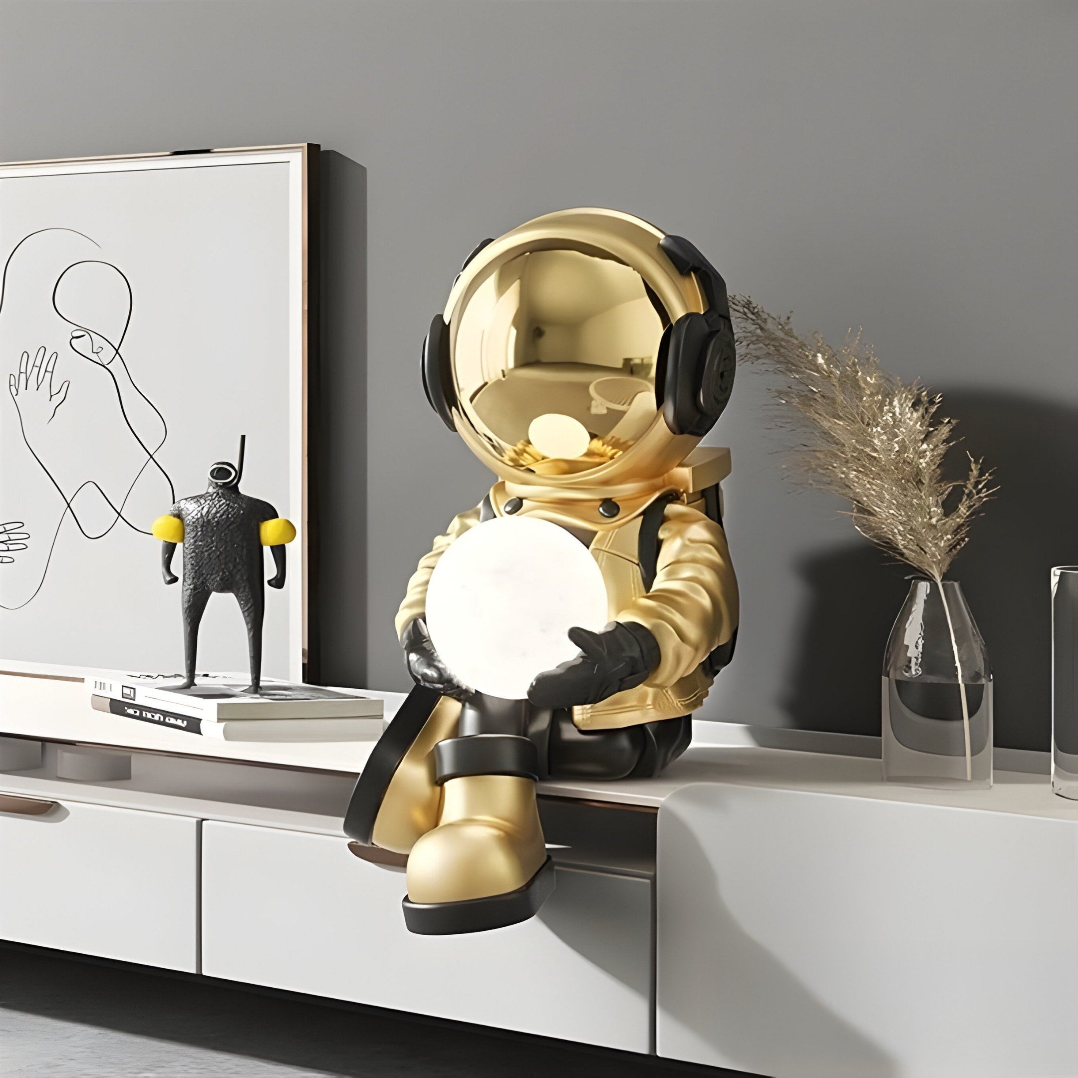 The Celestial Gold Astronaut Glow Seated I Sculpture by Giant Sculptures sits on a sleek white cabinet, holding a white orb. Its paired with minimalist art and a black abstract sculpture. A vase of dried grass adds texture against the gray backdrop, completing this celestial decor.