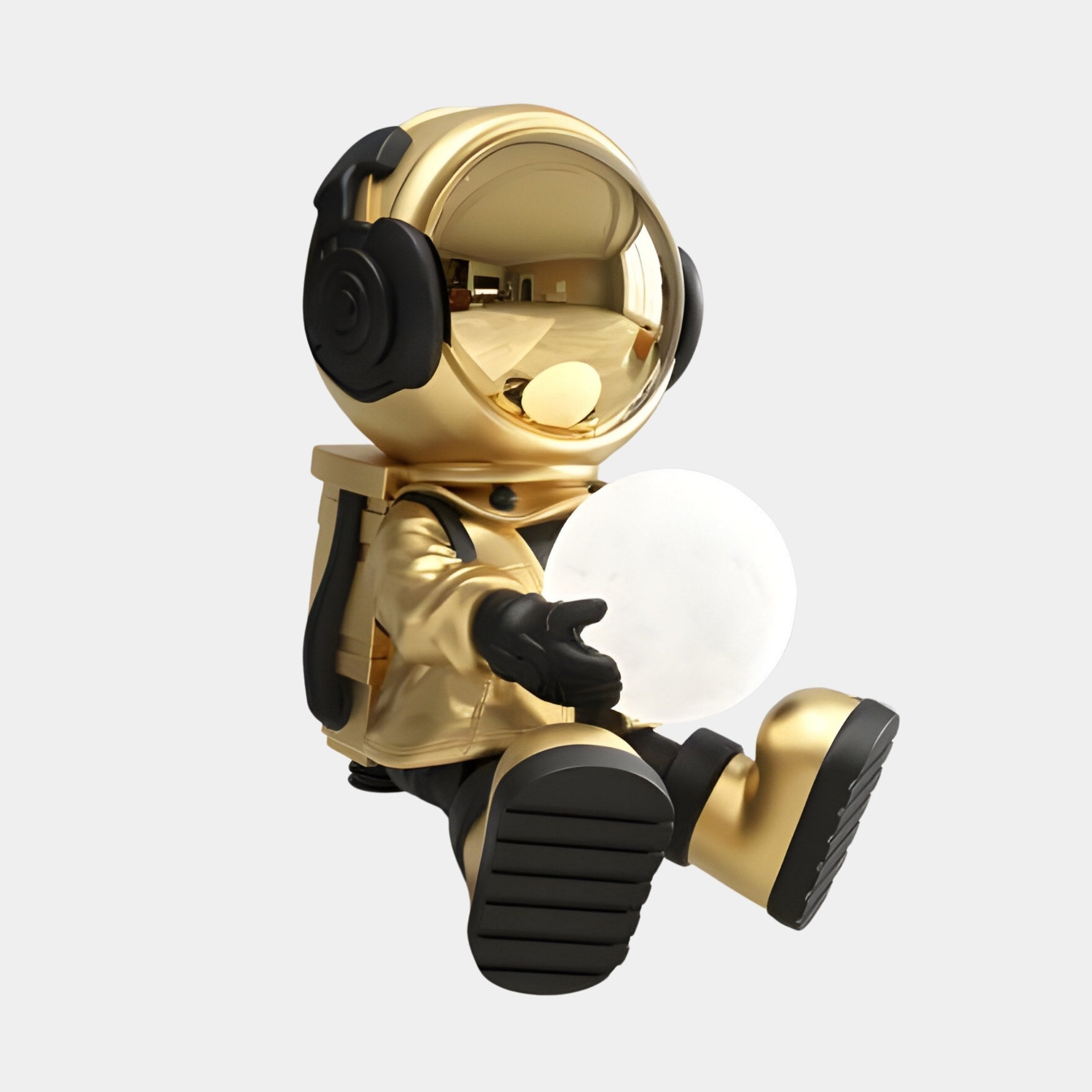 The Celestial Gold Astronaut Glow Seated II Sculpture by Giant Sculptures features a 53cm golden astronaut with headphones, cradling a glowing orb against a plain background. Its reflective helmet mirrors its surroundings, capturing the essence of a space-inspired lamp.