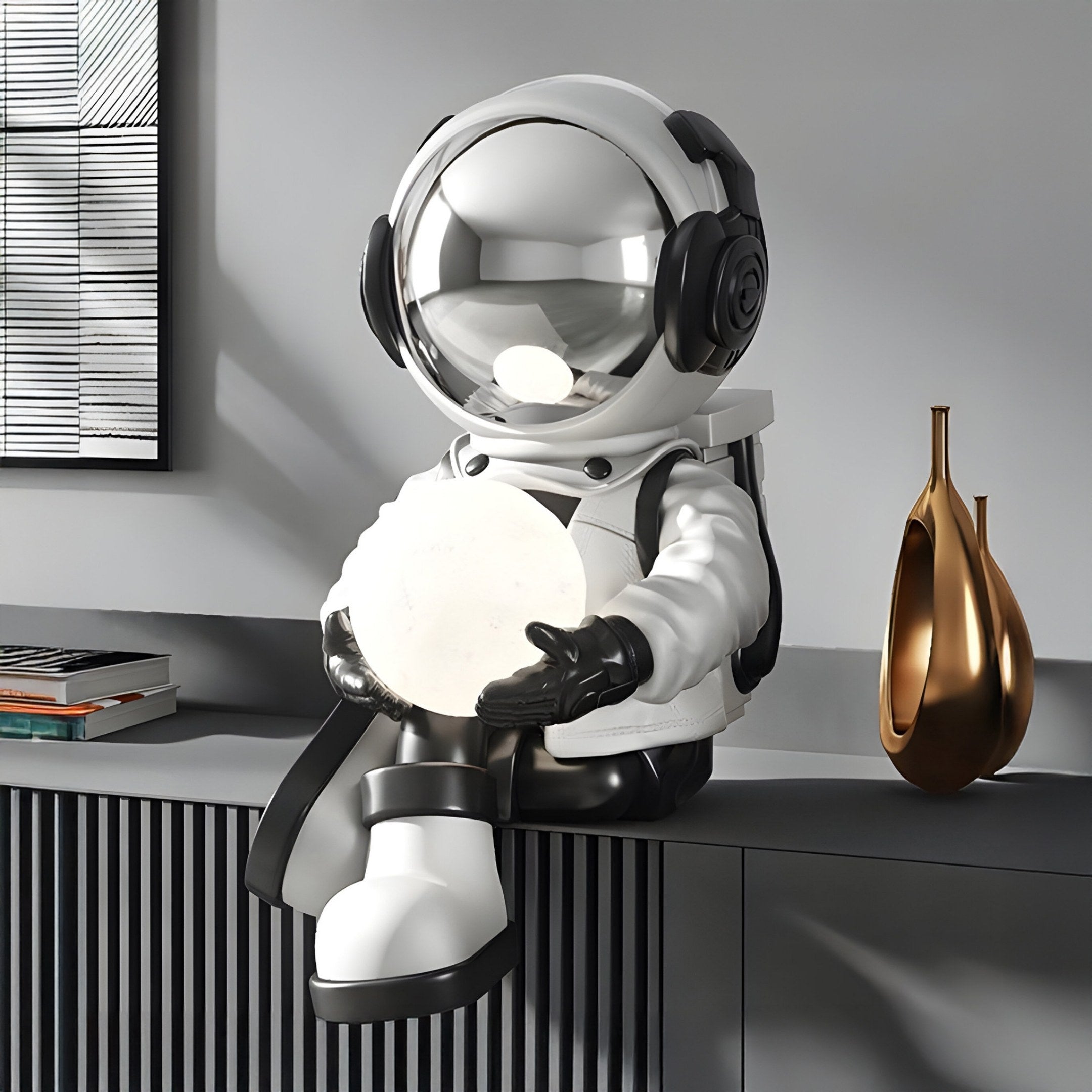 A Lunar Silver Astronaut Glow Seated I Sculpture by Giant Sculptures, sits with headphones on a gray shelf holding a white object. A copper vase and books complete the modern look. It features USB charging and adjustable lighting colors for convenience. Dimensions: 53cm.