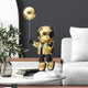 On a round marble table, the Celestial Gold Astronaut Balloon Sculpture - 55cm by Giant Sculptures rests against a backdrop of a white sofa, black wall, and potted plant, accompanied by stacked books.