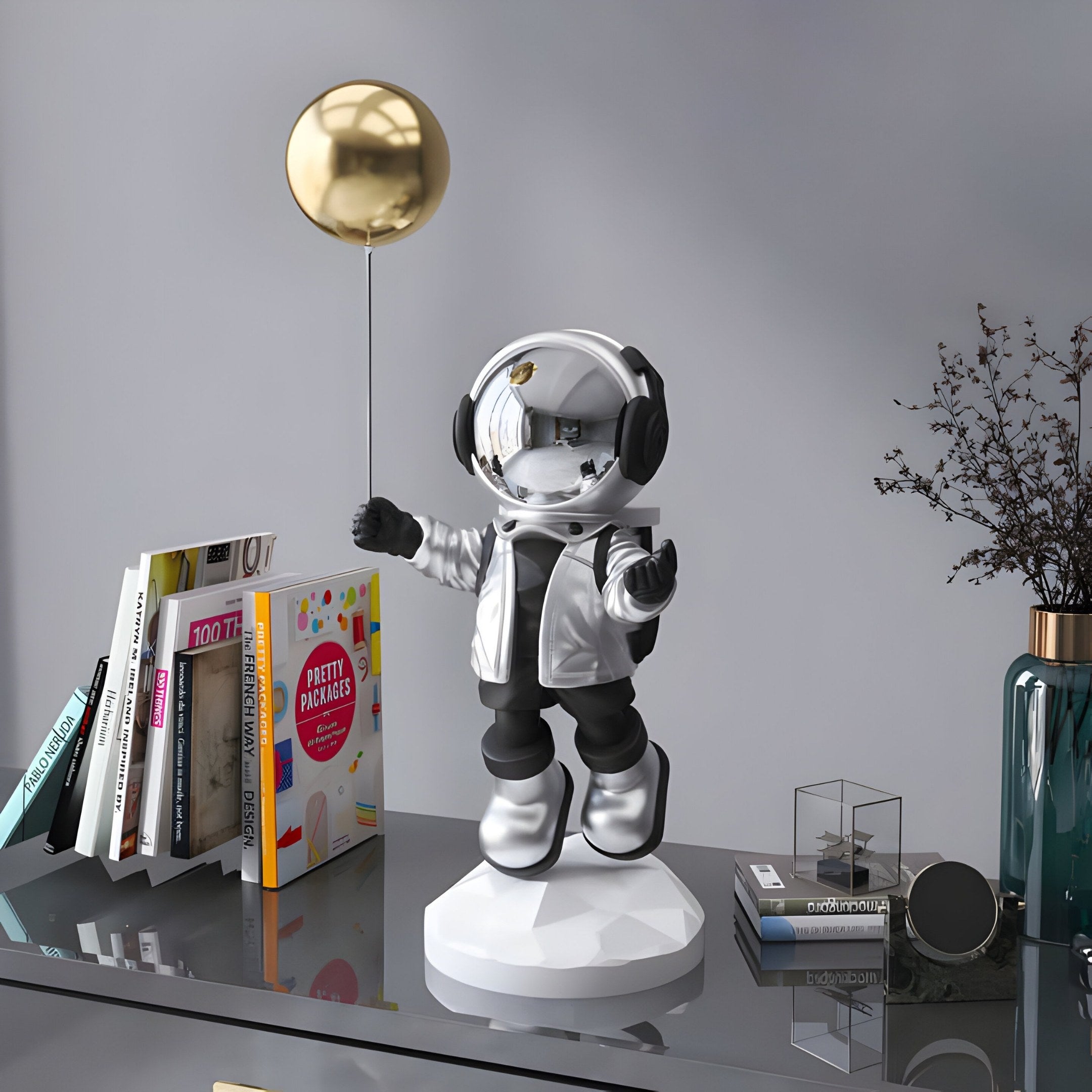 The Giant Sculptures Lunar Silver Astronaut Balloon Sculpture - 55cm stands on a reflective surface, complete with a shiny gold balloon and surrounded by books, a potted plant, and decorative items, creating a stylish cosmic theme in the modern display.