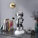 The Giant Sculptures Lunar Silver Astronaut Balloon Sculpture - 55cm stands on a reflective surface, complete with a shiny gold balloon and surrounded by books, a potted plant, and decorative items, creating a stylish cosmic theme in the modern display.