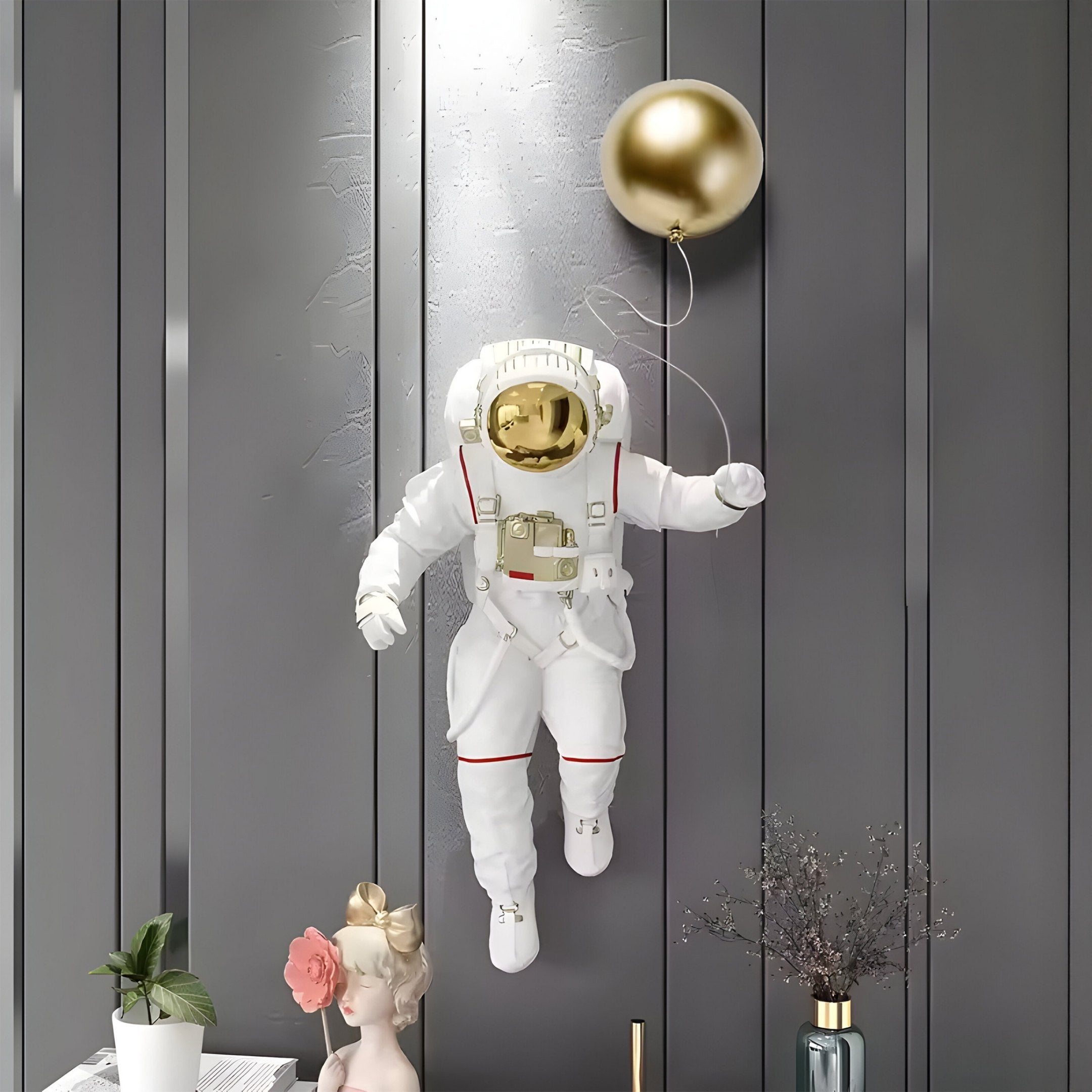 The White 3D Cosmic Lift Balloon Astronaut Wall Art by Giant Sculptures, featuring a gold visor, elegantly floats in front of a dark paneled wall. Nearby, a vase with branches and a classical bust with a gold crown and flowers enhance the decorative sculpture display.