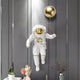 The White 3D Cosmic Lift Balloon Astronaut Wall Art by Giant Sculptures, featuring a gold visor, elegantly floats in front of a dark paneled wall. Nearby, a vase with branches and a classical bust with a gold crown and flowers enhance the decorative sculpture display.