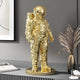 A Gilded Gold Space Astronaut Standing Sculpture by Giant Sculptures enhances a modern table. Set against a gray wall, abstract art, and plants, it creates an interstellar decor vibe in this sophisticated contemporary setting.