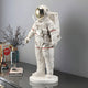 A Cloud White Space Astronaut Standing Sculpture - 58cm by Giant Sculptures, with a reflective gold visor, is displayed on a table featuring vases of dried flowers, a glass cuboid, and colorful books. This space-themed décor contrasts elegantly against the plain gray wall.