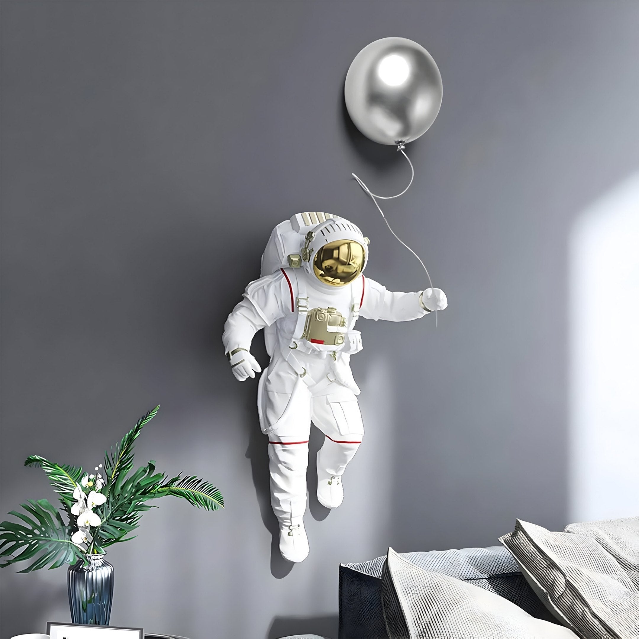 The White 3D Cosmic Lift Silver Balloon Astronaut Wall Art by Giant Sculptures showcases a metallic helmet visor set against a gray backdrop. On the table, a vase with green leaves and white flowers enhances this cosmic scene.