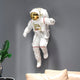 The White 3D Cosmic Lift Astronaut Wall Art (58cm) by Giant Sculptures features an astronaut in a white suit with a gold visor floating in front of a gray wall. Below, a green plant and part of a beige couch provide contrast, boosting the cosmic charm of this ethereal scene.