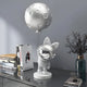 A sleek, modern room showcases the 60cm Cosmic Chrome Astronaut Bunny Moon Balloon Sculpture by Giant Sculptures on a tall stand. Nearby, books and a glass vase with dry twigs rest on a minimalist, reflective storage box that complements the cosmic decor.