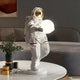 The Giant Sculptures White Space Astronaut Glow Orb Sculpture (60cm) with adjustable lighting colors stands out with its reflective gold visor and red-accented white spacesuit on a living room table, complemented by a curved sofa and abstract wall art in the background.