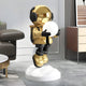 The Celestial Gold Astronaut Glow Hover Sculpture by Giant Sculptures stands on a white platform in a modern room. Featuring a reflective helmet and illuminating orb, the astronaut in a gold jacket, black gloves, boots, and headphones is perfectly placed by the sofa as a touch switch lighting centerpiece.