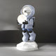 The Cosmic Sky Astronaut Glow Hover Sculpture by Giant Sculptures is a 60cm figure in a white suit and helmet, holding a glowing moon-like object. It stands on a faceted white base set against a dark gray background, resembling an exquisite hover sculpture.