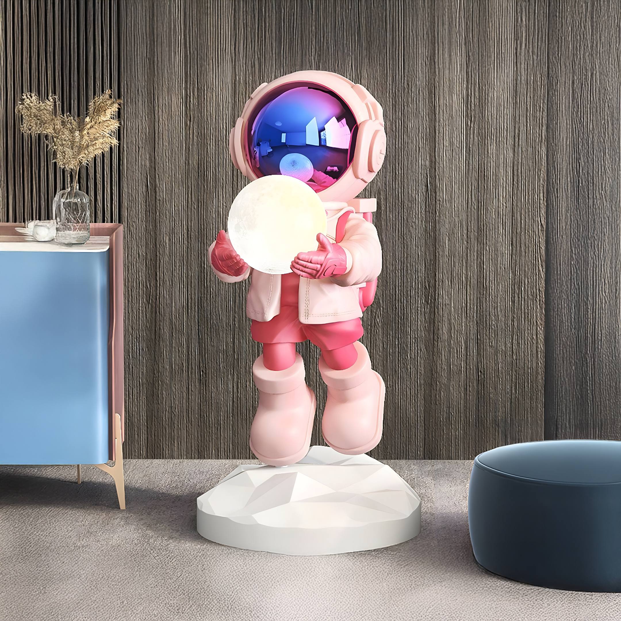 The Stellar Pink Astronaut Glow Hover Sculpture by Giant Sculptures beams brightly indoors on its geometric pedestal, reflecting ambient light against a textured wall beside a blue cabinet with dried flowers and a round blue ottoman.