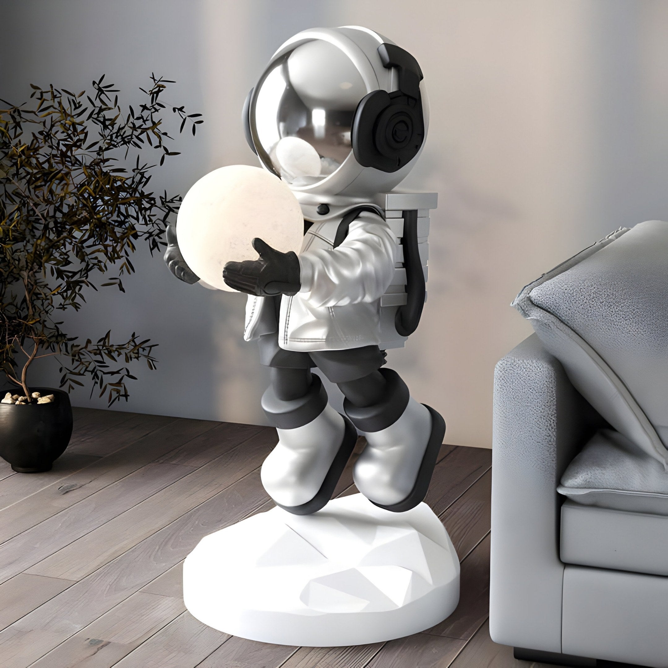 The Lunar Silver Astronaut Glow Hover Sculpture by Giant Sculptures, in a sleek lunar silver suit, holds a glowing orb on a geometric white base. Its displayed on a wooden floor near a gray sofa, with a plant behind, creating a cozy modern scene.