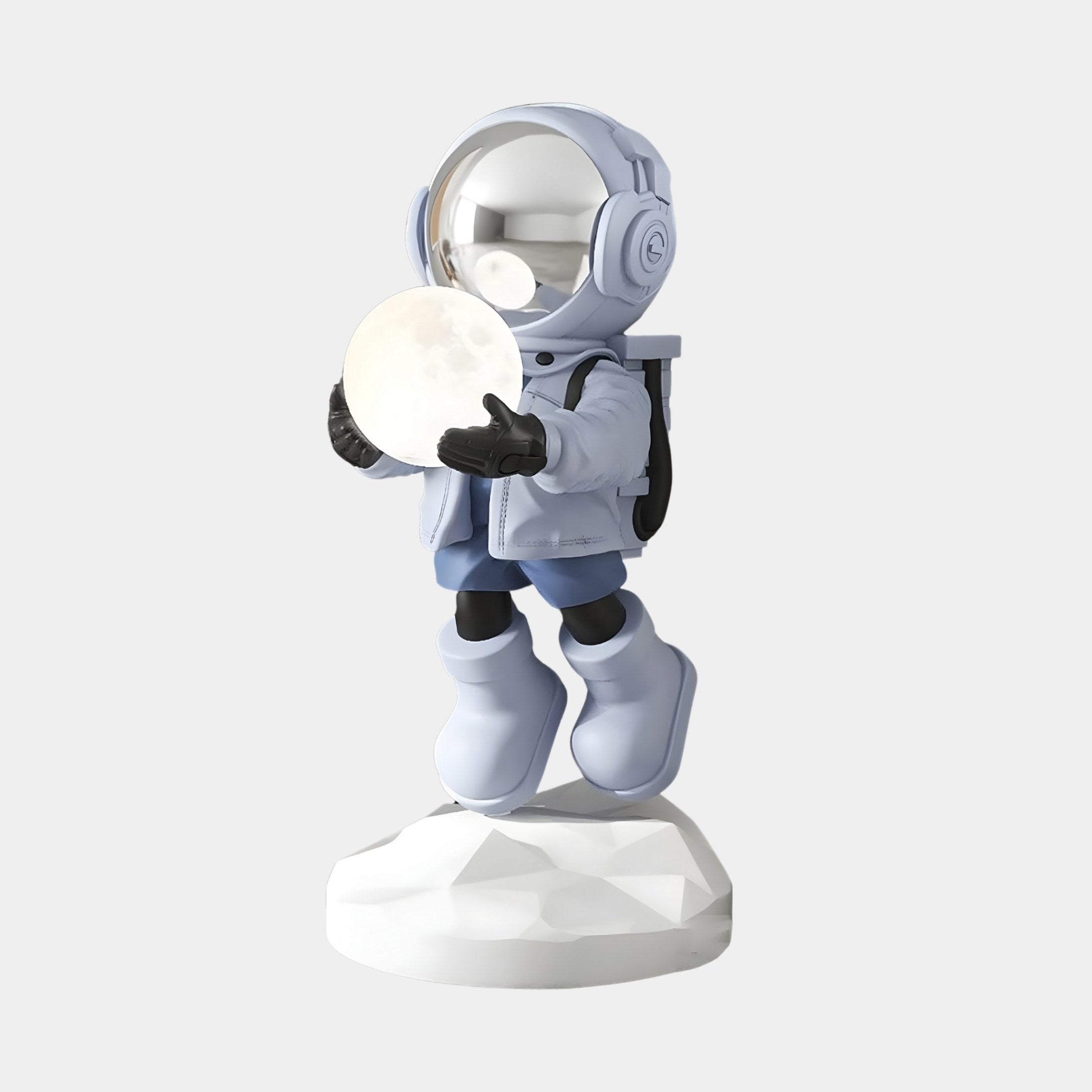 The Giant Sculptures Cosmic Sky Astronaut Glow Hover Sculpture - 60cm features a small astronaut in a light blue suit holding a glowing moon, standing on a white rocky base. It has a reflective helmet, black gloves and boots, and adds celestial charm to any plain gray backdrop.