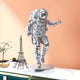 The Cosmic Chrome Space Astronaut Explorer Sculpture by Giant Sculptures, featuring a reflective silver finish, is placed on a white surface beside an Eiffel Tower wireframe. A peach-colored wall and a green cabinet with a small globe and plant are partially visible in the background.