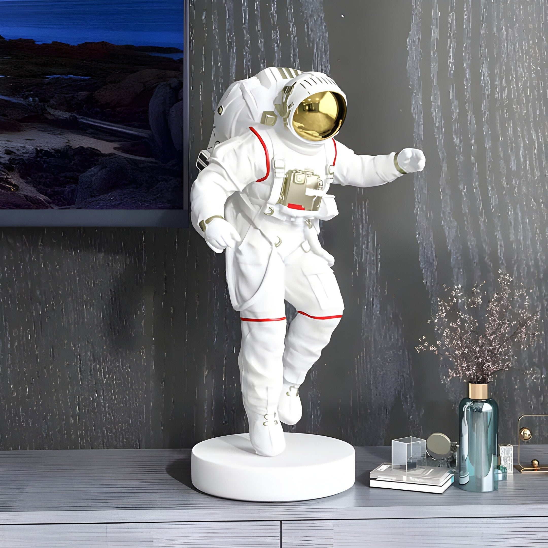 The White Space Astronaut Explorer Sculpture by Giant Sculptures stands 63cm tall with a golden visor, white suit, and round base. Positioned on a gray surface and textured wall backdrop, its complemented by a small plant, glass bottles, and picture frame for a complete space-themed decor setup.